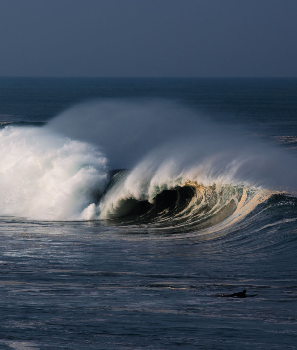 Large ocean wave crashing with foam and surfer near shore.