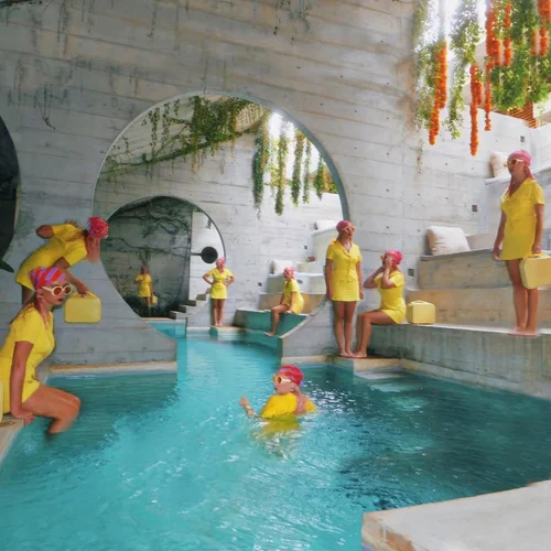 Children in yellow swimwear play in an indoor pool beside fountain-like arch.