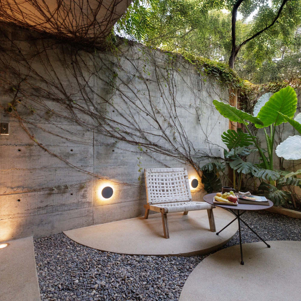 Patio con pared de concreto y silla de mimbre, plantas tropicales y lámparas cálidas