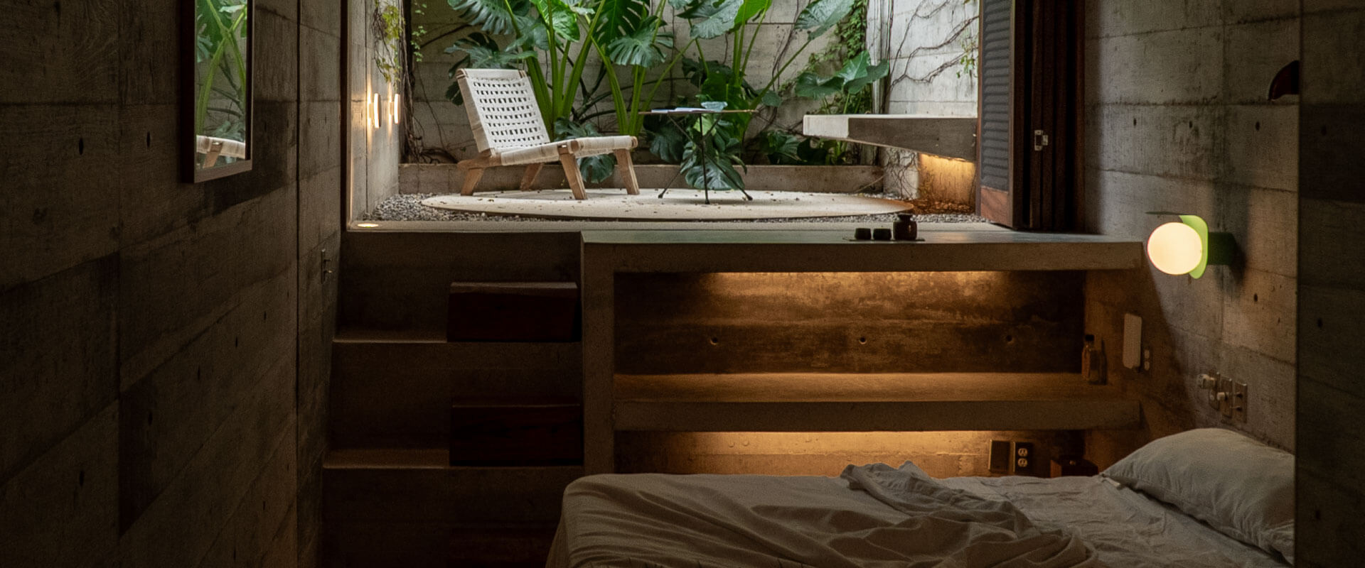 Modern bedroom with small window, concrete walls, lush plants, and a bed