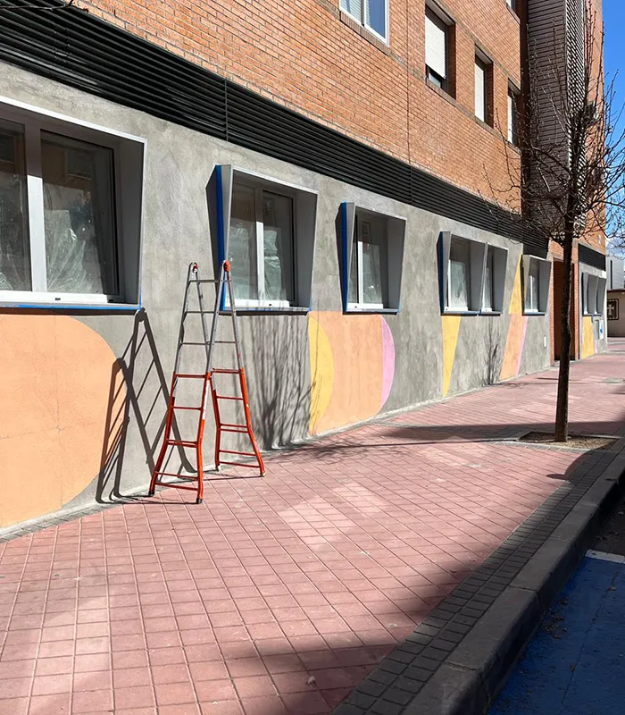 Escalera roja apoyada en una pared con ventanas y mural de formas geométricas en un día soleado.