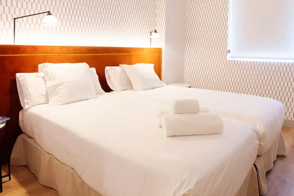 Dos camas individuales unidas con sábanas blancas, almohadas y toallas dobladas, lámparas y fondo de papel tapiz geométrico.