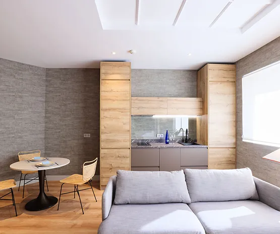 Sala de estar moderna con sofá gris, cocina compacta y mesa redonda con sillas en madera clara.
