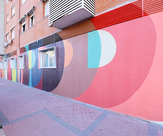 Mural colorido con formas geométricas sobre la pared de un edificio junto a una acera. Ventanas alineadas a la izquierda.