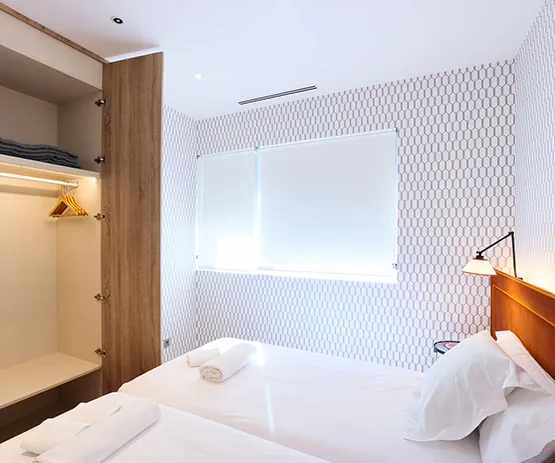 Habitación minimalista con dos camas, pared con diseño hexagonal, ventana con persiana y armario abierto con perchas.