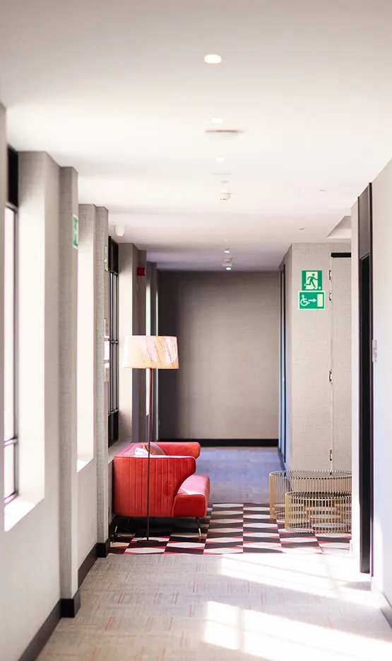 Pasillo iluminado con lámpara, sillón rojo y señal de salida a la derecha.