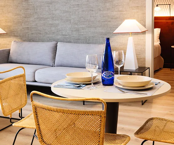 Sala moderna con sofá gris, mesa redonda con platos, copas y botella azul, sillas de mimbre y lámparas de mesa.