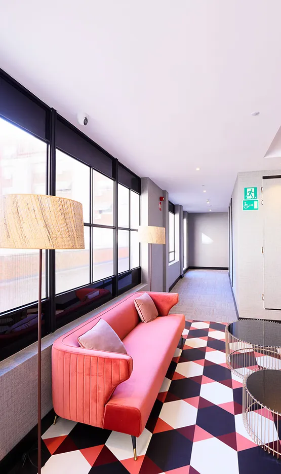 Sala moderna con sofá rosa, cojines, alfombra geométrica roja y blanca, y lámparas a ambos lados, junto a ventanas grandes.