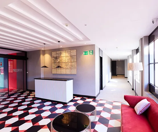 Vestíbulo moderno con piso geométrico. Muebles en rojo y negro, recepción blanca y luces colgantes. Iluminación natural.