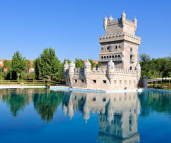 Torre medieval de piedra con reflejo en el agua, rodeada de árboles y cielo azul.