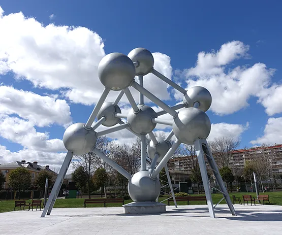 Escultura metálica con forma de átomo gigante bajo un cielo azul con nubes esponjosas.