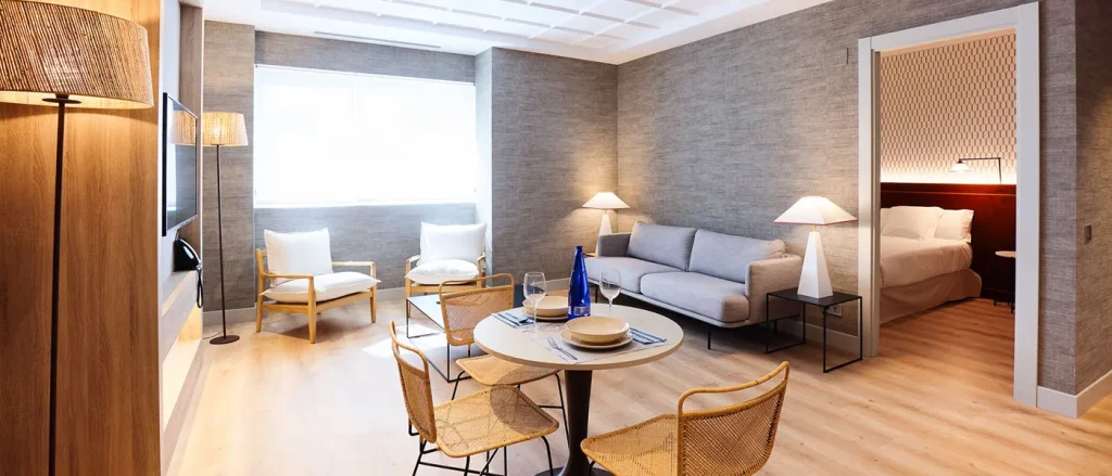 Sala moderna con sofá gris, mesa redonda para comer, lámparas de pie y acceso a habitación con cama. Decoración minimalista.