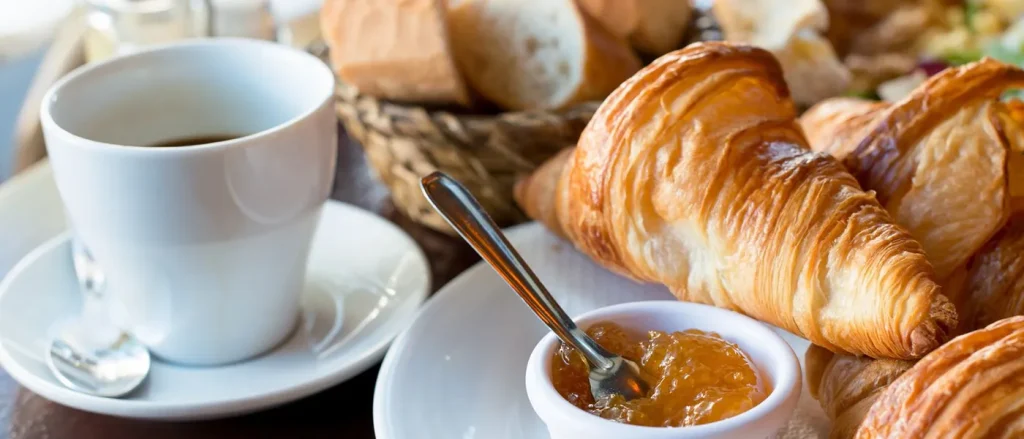 Croissant con mermelada y café en taza blanca sobre mesa de desayuno. Fondo con pan y cesta de mimbre.
