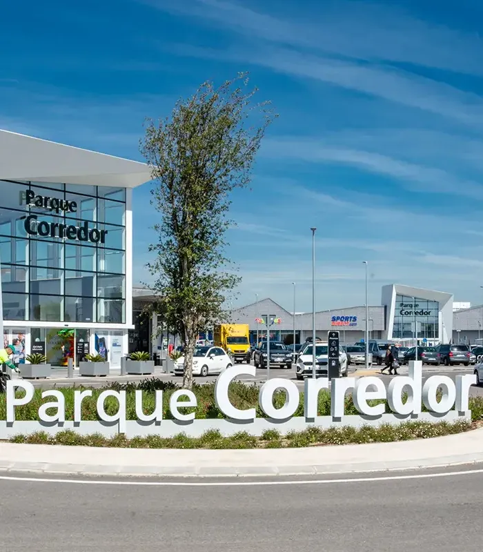 Centro comercial "Parque Corredor" con coches aparcados y cielo despejado. Árboles y vegetación al frente.