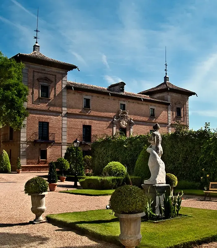 Edificio histórico de ladrillo con torres, rodeado de jardines verdes, con una estatua en primer plano bajo el cielo azul.