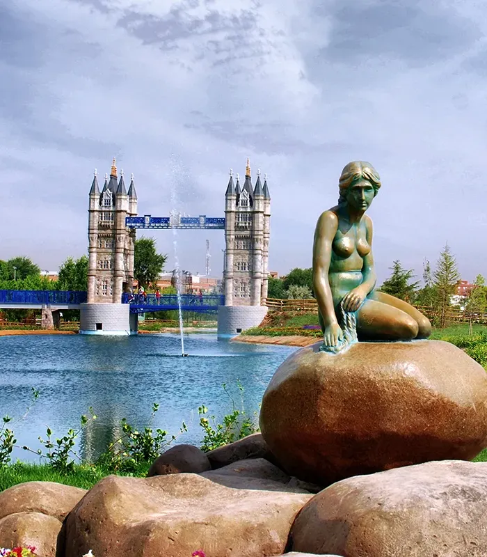 Una estatua sobre una roca frente a un puente de estilo gótico junto a un lago con fuente y árboles alrededor.