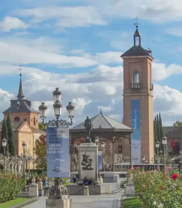 Plaza con estatua, quiosco y torre. Cielo azul con nubes. Faroles antiguos y vegetación alrededor.