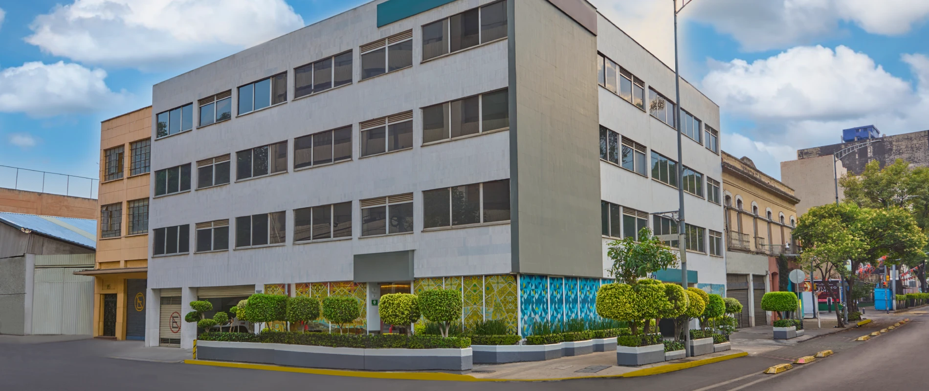 Edificio de varios pisos en esquina, con ventanas y plantas al frente, bajo cielo azul y nubes.