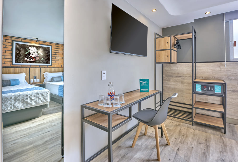 Habitaci&oacute;n moderna de hotel con cama, escritorio, televisor, caja fuerte y espejo.