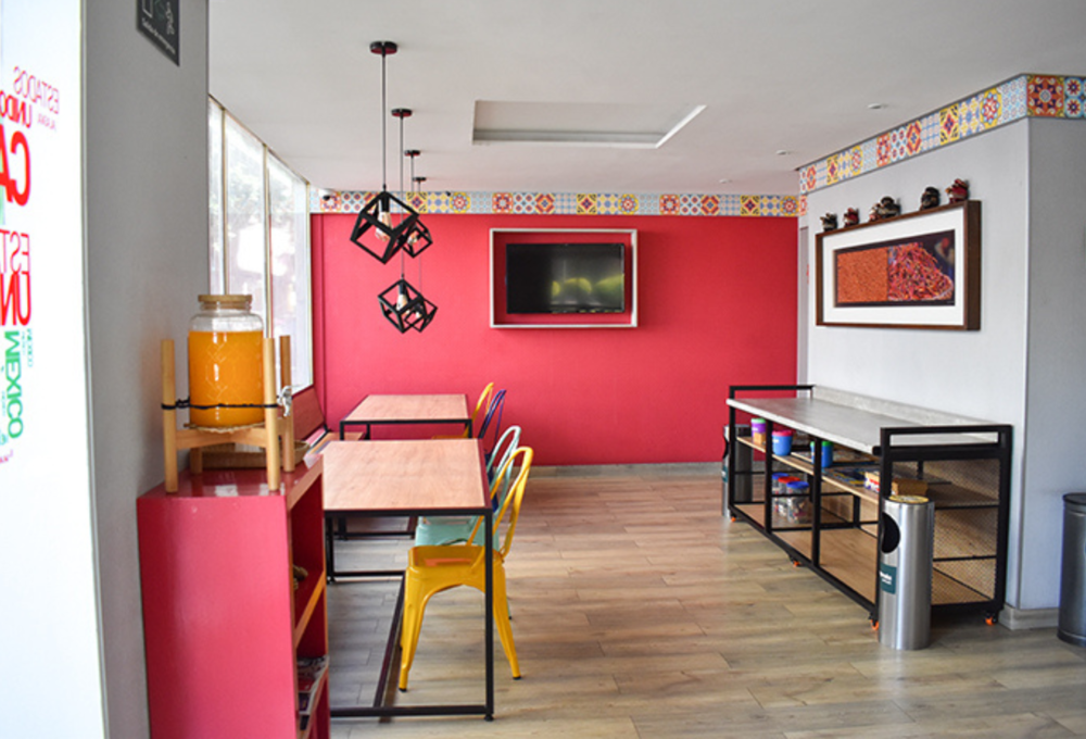 Sala colorida con mesas, sillas amarillas y TV en la pared. Bebida en jarra sobre estante rojo.