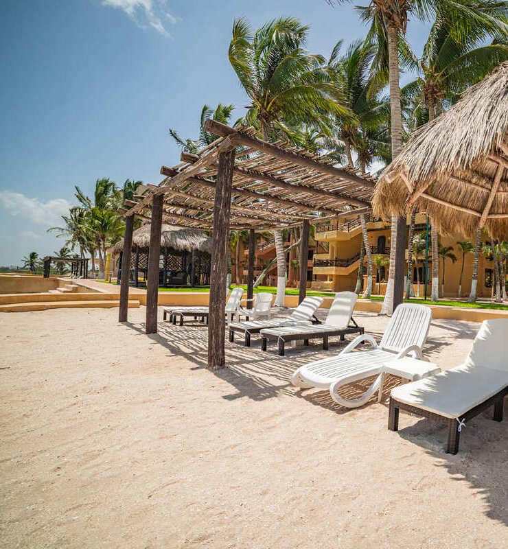 Tumbonas blancas bajo una pérgola de madera en una playa de arena, rodeada de palmeras y edificios al fondo.