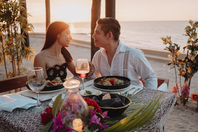 Pareja disfrutando de una cena romántica al atardecer en la playa con comida y vino sobre la mesa decorada.