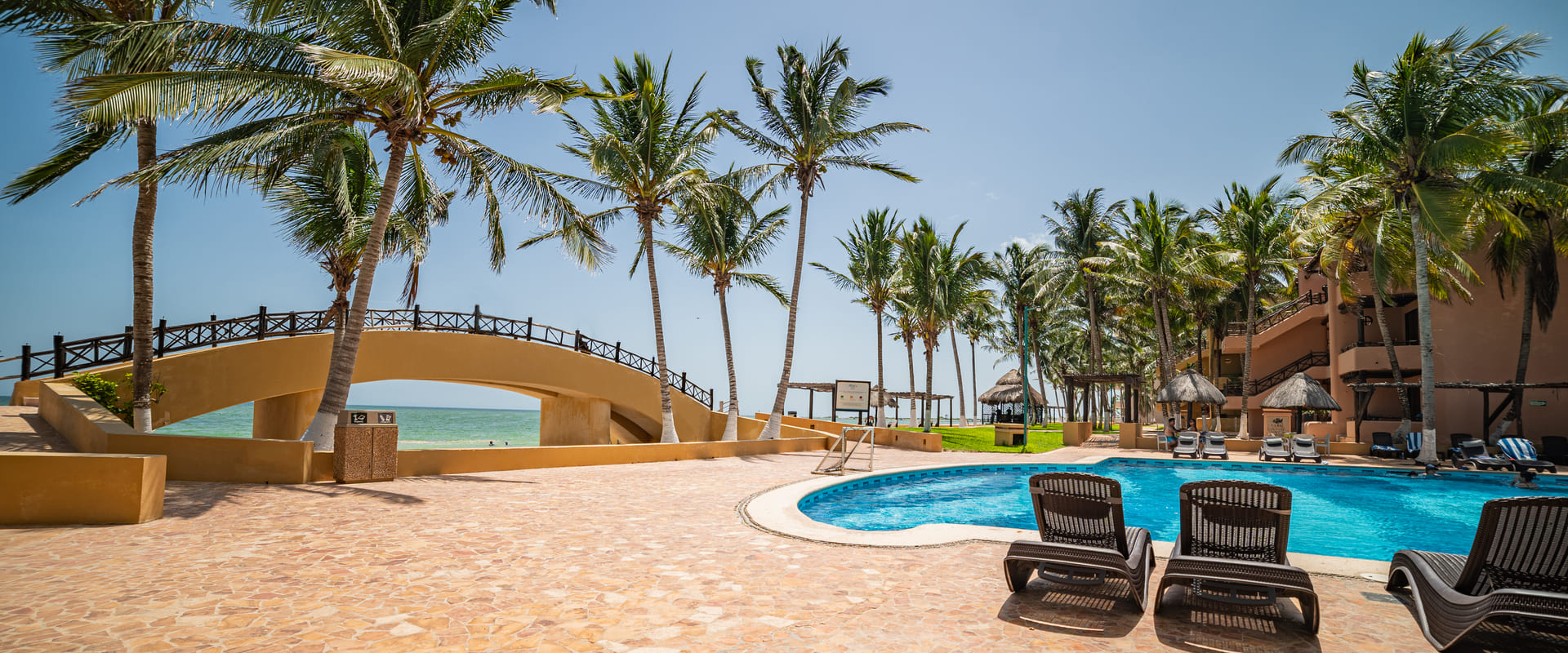 Piscina con sillones, palmeras y puente en un resort junto al mar bajo un cielo despejado.