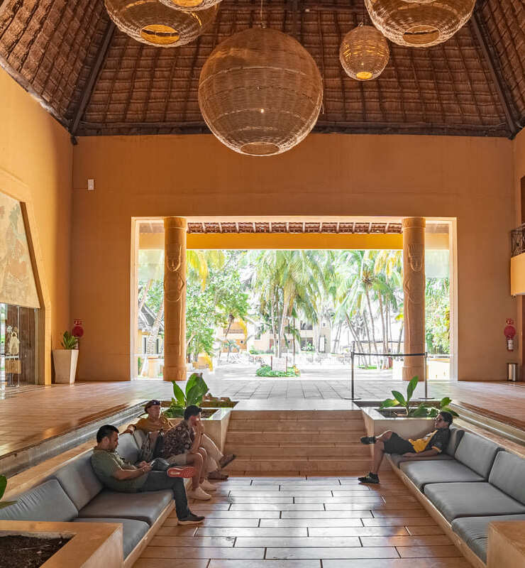 Sala con techo de paja, lámparas colgantes, sofás grises, y personas sentadas; vista de palmeras al fondo.