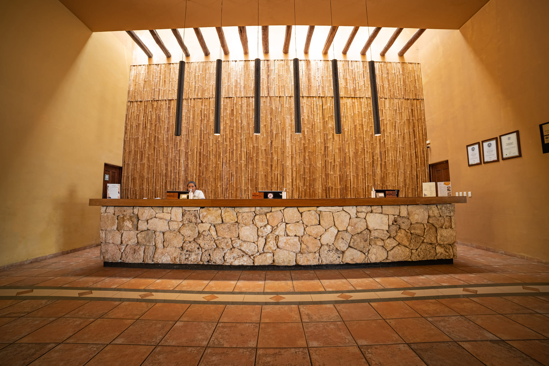 Recepción de hotel moderna con mostrador de piedra, fondo de bambú y luces colgantes, atendida por un recepcionista.