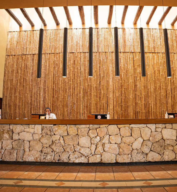 Mostrador de recepción de hotel con pared de bambú, lámparas colgantes y base de piedra. Persona trabajando detrás.