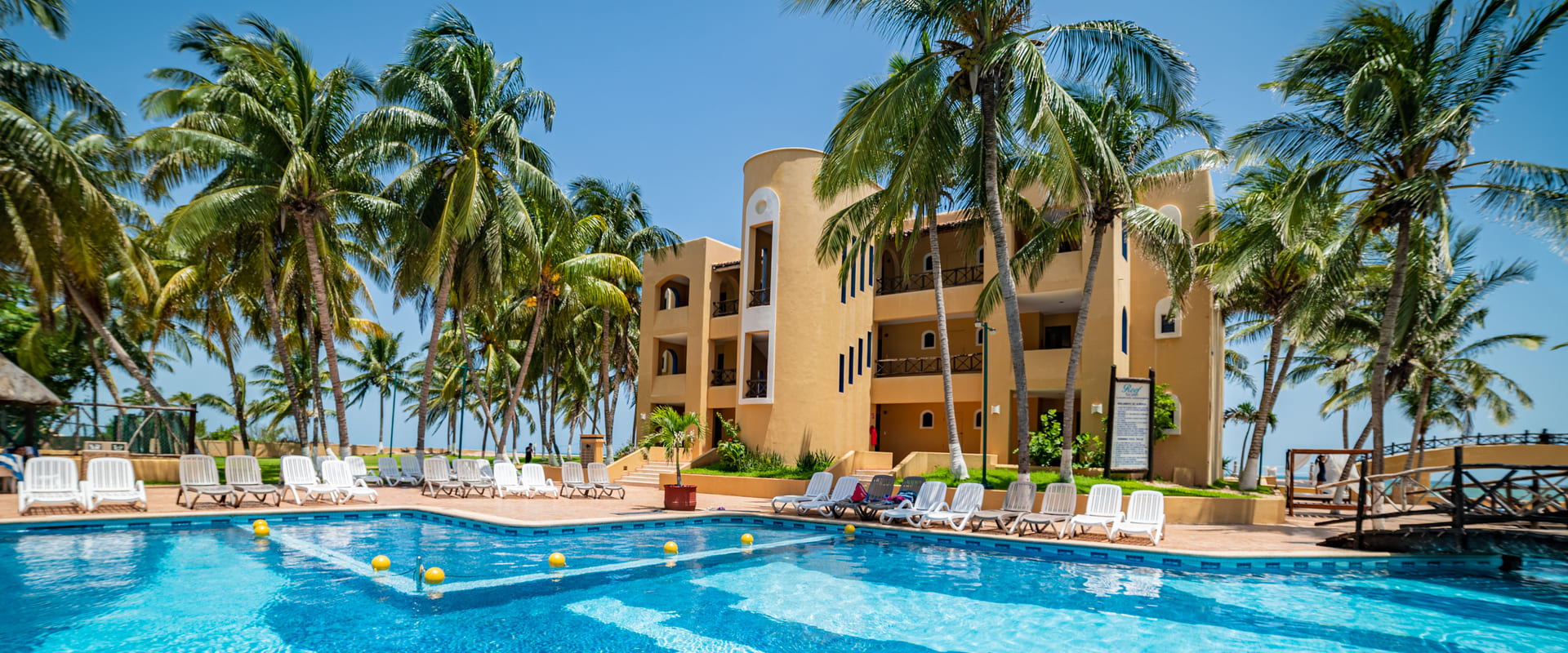 Piscina frente a un hotel de tres pisos rodeado de palmeras bajo un cielo azul claro.
