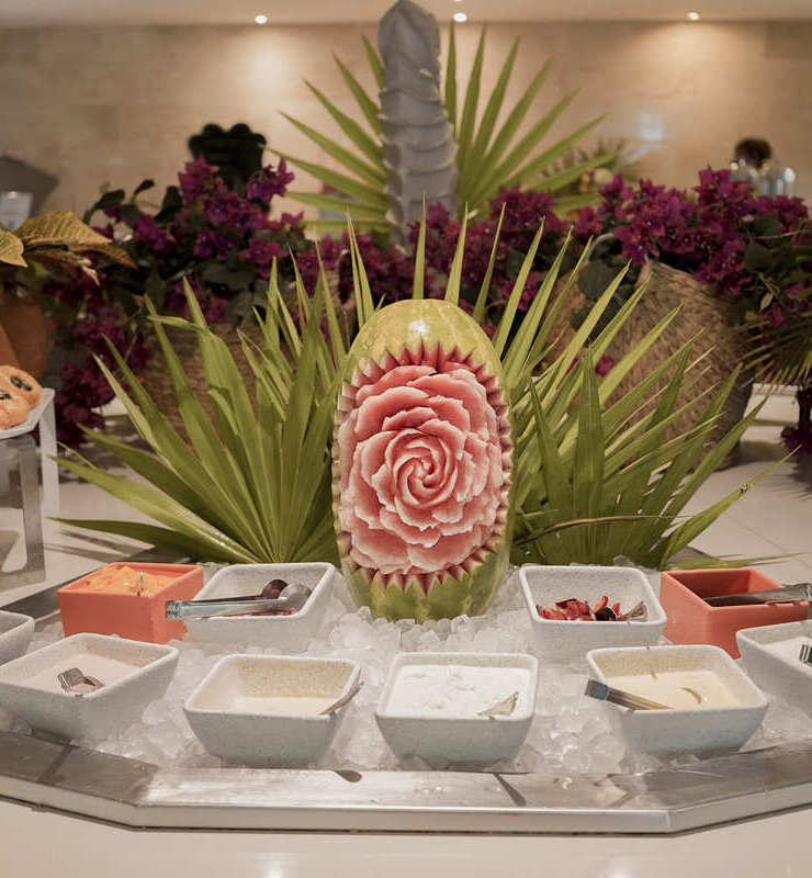 Decoración de buffet con sandía tallada en forma de flor y diversos cuencos con condimentos sobre hielo.