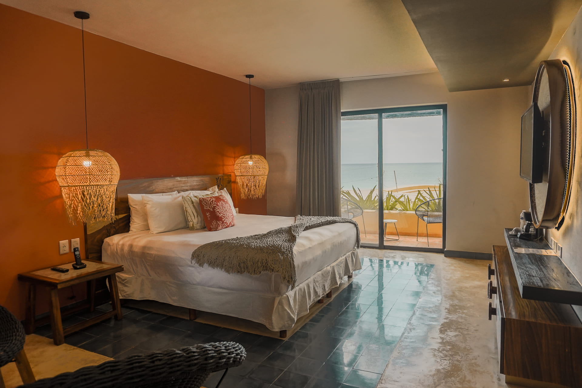 Habitación moderna con cama doble, luces colgantes y vista al mar desde el balcón, decorada en tonos cálidos.