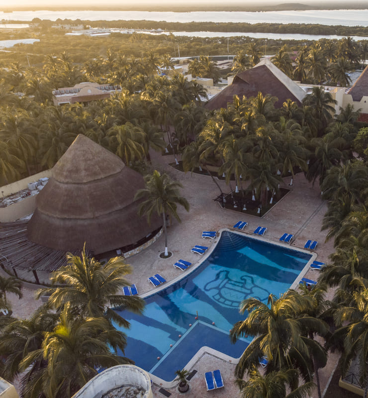 Vista aérea de una piscina rodeada de palmeras, con edificios de techo de paja y hamacas azules, al atardecer.