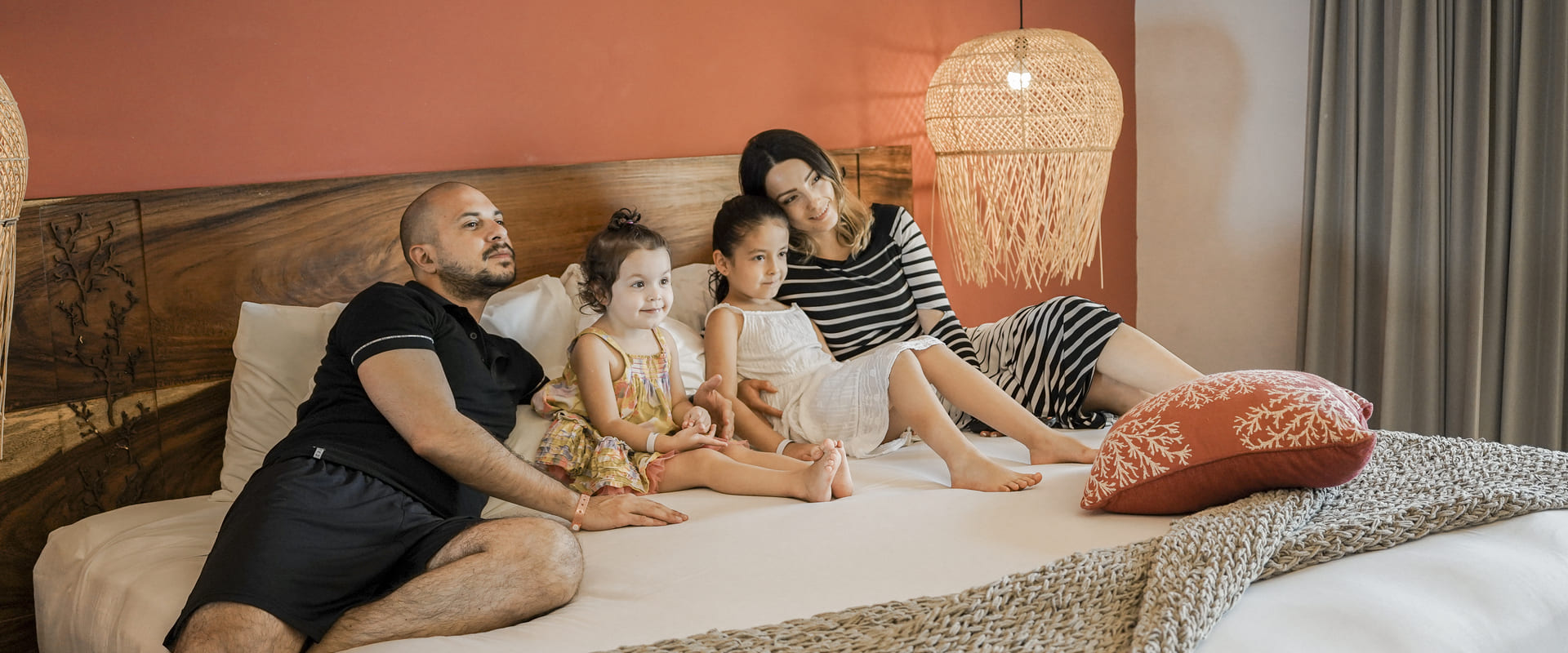 Familia sonríe y descansa en una cama grande con lámpara colgante y decoración acogedora.