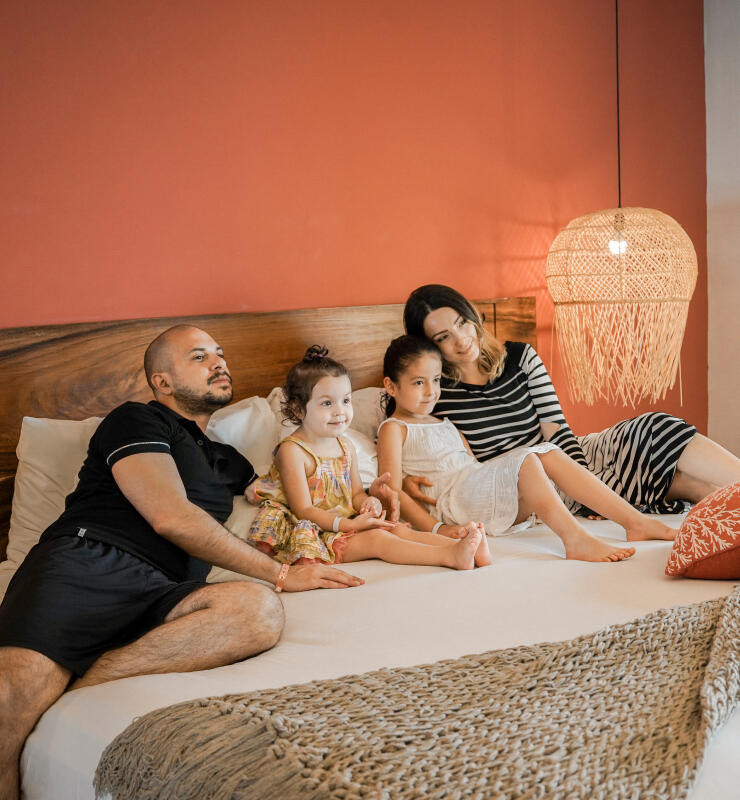 Familia sentada en una cama grande con un fondo de pared roja y una lámpara colgante de mimbre al lado.