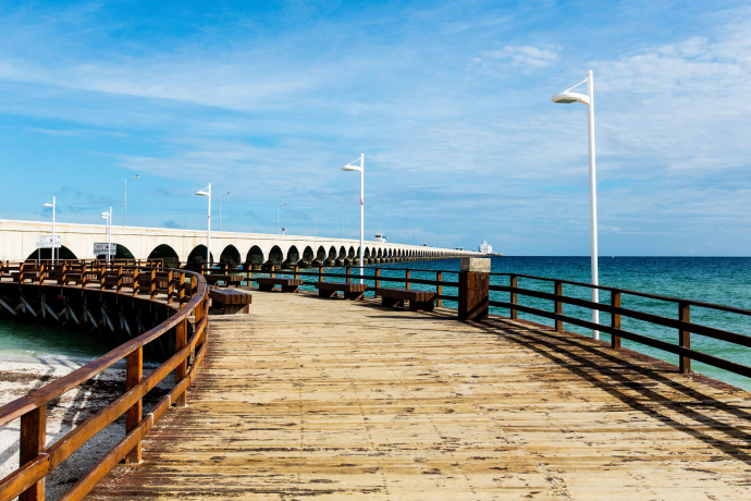Muelle de madera junto al mar azul, con farolas blancas y un cielo parcialmente nublado.