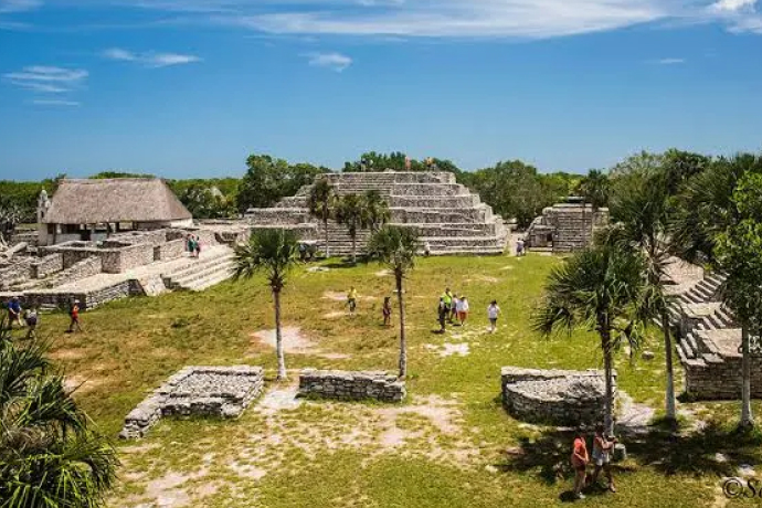 Ruinas mayas en un paisaje verde con turistas caminando, rodeadas de estructuras de piedra y árboles bajo un cielo azul.