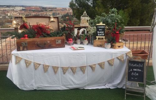 Mesa decorada con tema rústico navideño, con maleta, plantas y pizarra que dice "Bienvenidos a nuestra boda".