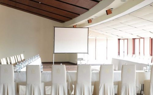 Sala de conferencias vacía con sillas blancas, mesa, botellas de agua y pantalla de proyección al fondo.