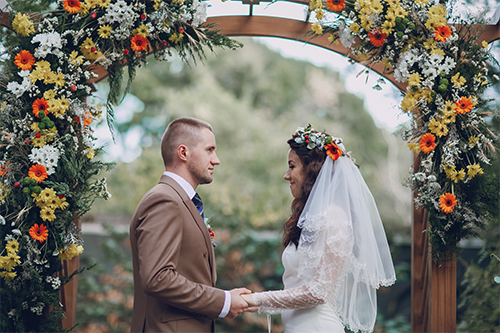Pareja de novios entrelazando manos bajo un arco floral en su boda al aire libre.