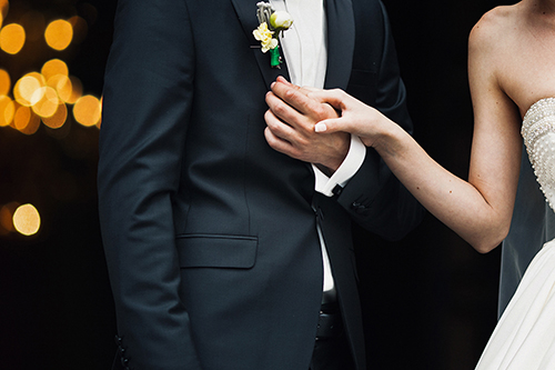 Persona con traje oscuro y persona con vestido blanco tomados de la mano, luces borrosas en el fondo.