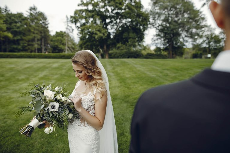 Novia en vestido blanco mira su ramo de flores en un jardín, con el novio de espaldas en primer plano.