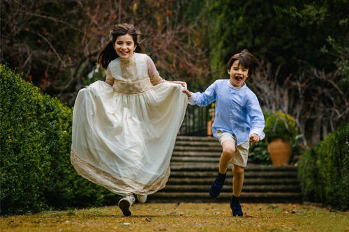 Niña y niño corren felices en un jardín, la niña lleva un vestido blanco y el niño una camisa azul con pantalones cortos.