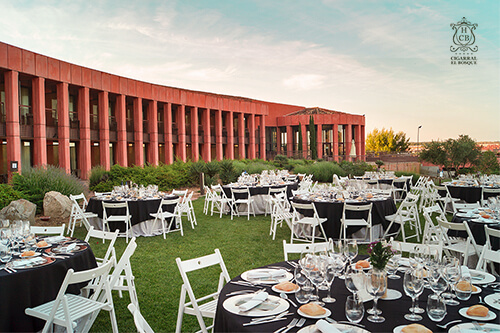 Evento al aire libre con mesas redondas y sillas blancas en un jardín, frente a un edificio de columnas rojas al atardecer.