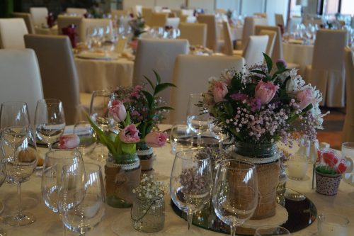 Mesas decoradas con flores rosadas y copas de cristal en un salón de eventos elegante y bien iluminado.