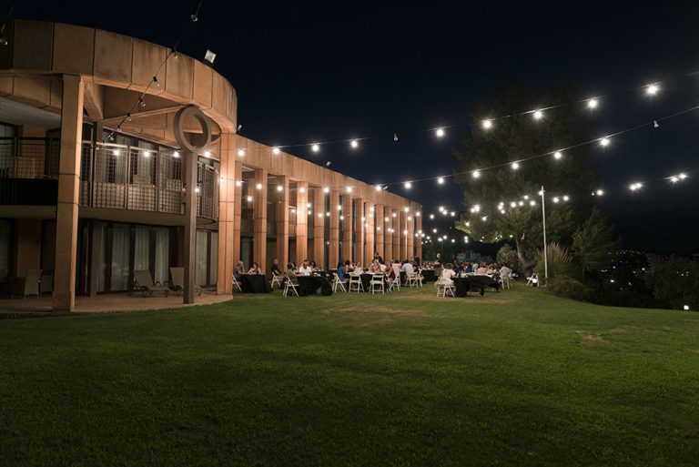 Evento nocturno al aire libre con luces colgantes y mesas en una terraza junto a un edificio iluminado.