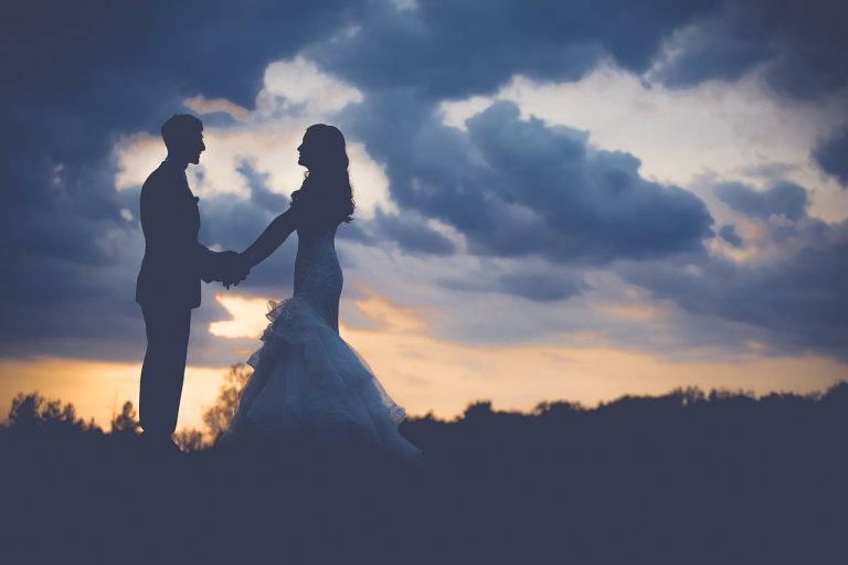 Silueta de pareja frente a cielo nublado y atardecer, tomados de la mano, él con traje y ella con vestido de novia.