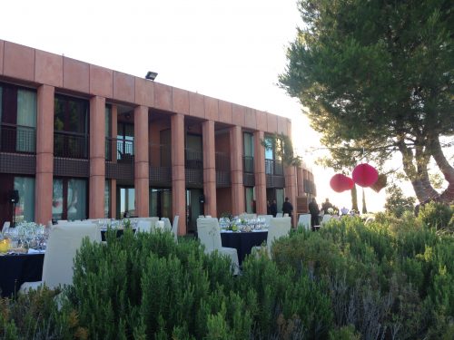 Edificio moderno con mesas al aire libre, decorado con globos rojos y rodeado de vegetación y árboles.