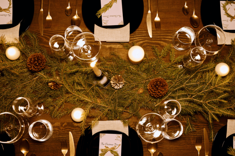 Mesa navideña decorada con ramas de pino, piñas, velas y copas de vino. Vajilla dispuesta elegantemente.